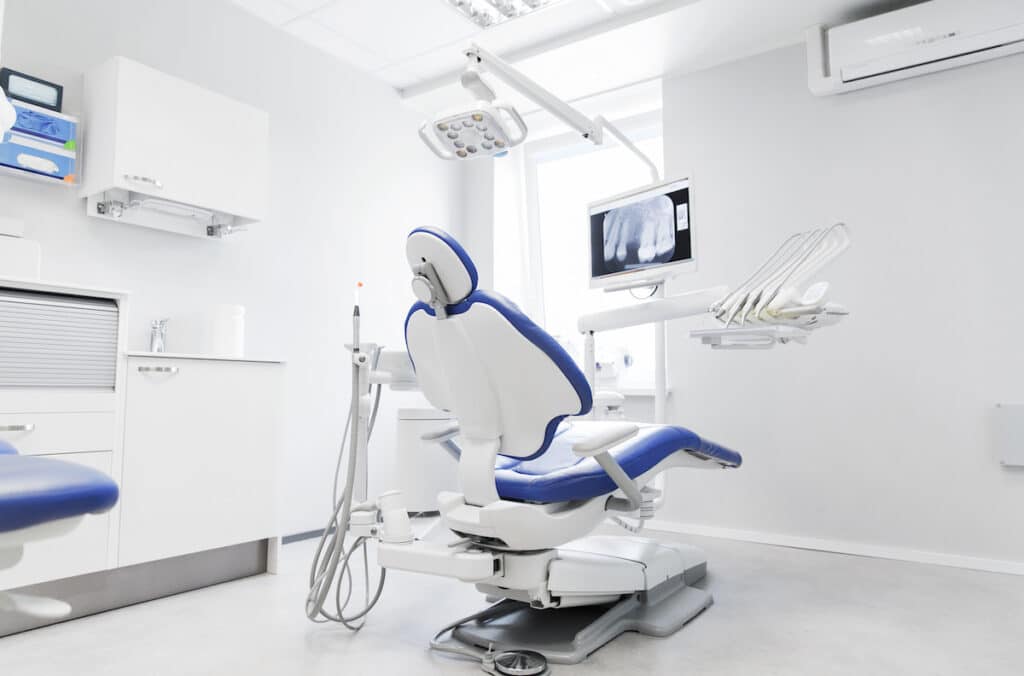 The interior of a bright, modern dental exam room. There is a dental chair facing a screen with a dental x-ray on it.