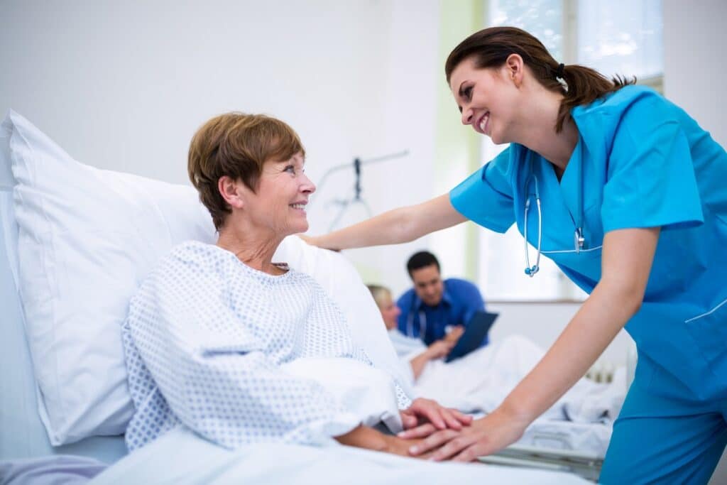 A nurse is leaning over an older woman in a hospital bed. They are both smiling.