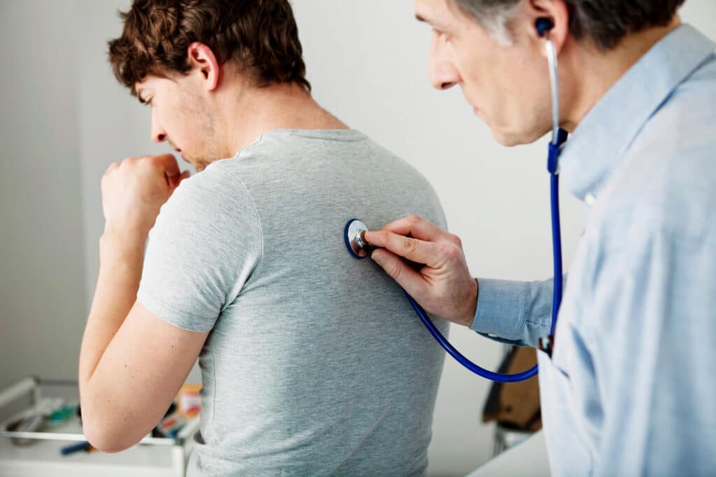 A medical professional holds his stethoscope to the back of a young adult male patient who is coughing.