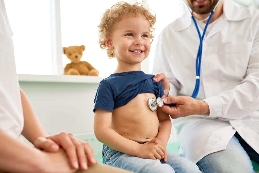 A little boy is smiling in an exam room. A male pediatrician is holding a stethoscope to his chest.