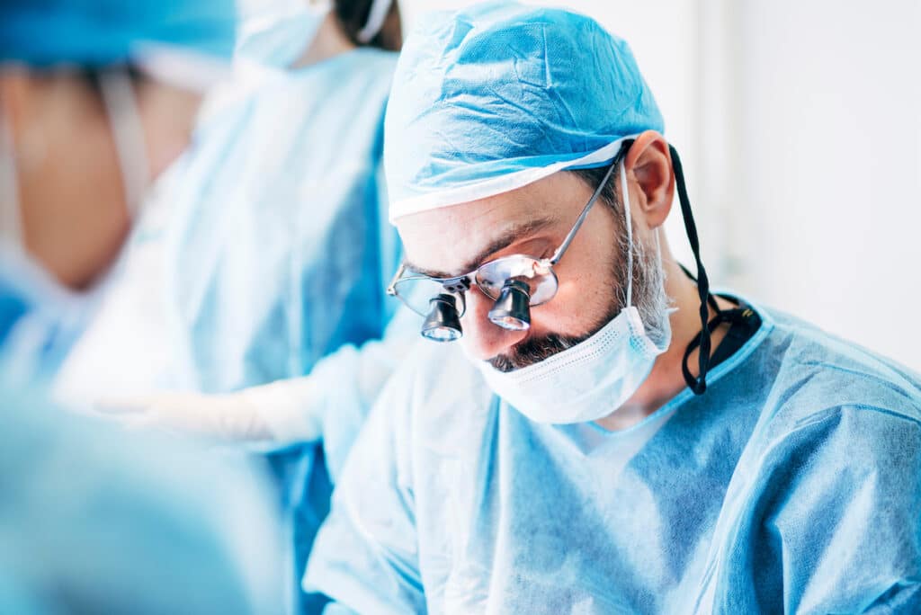 A close-up of a dentist in full scrubs in an operating room. There are two other dental professionals in the frame.