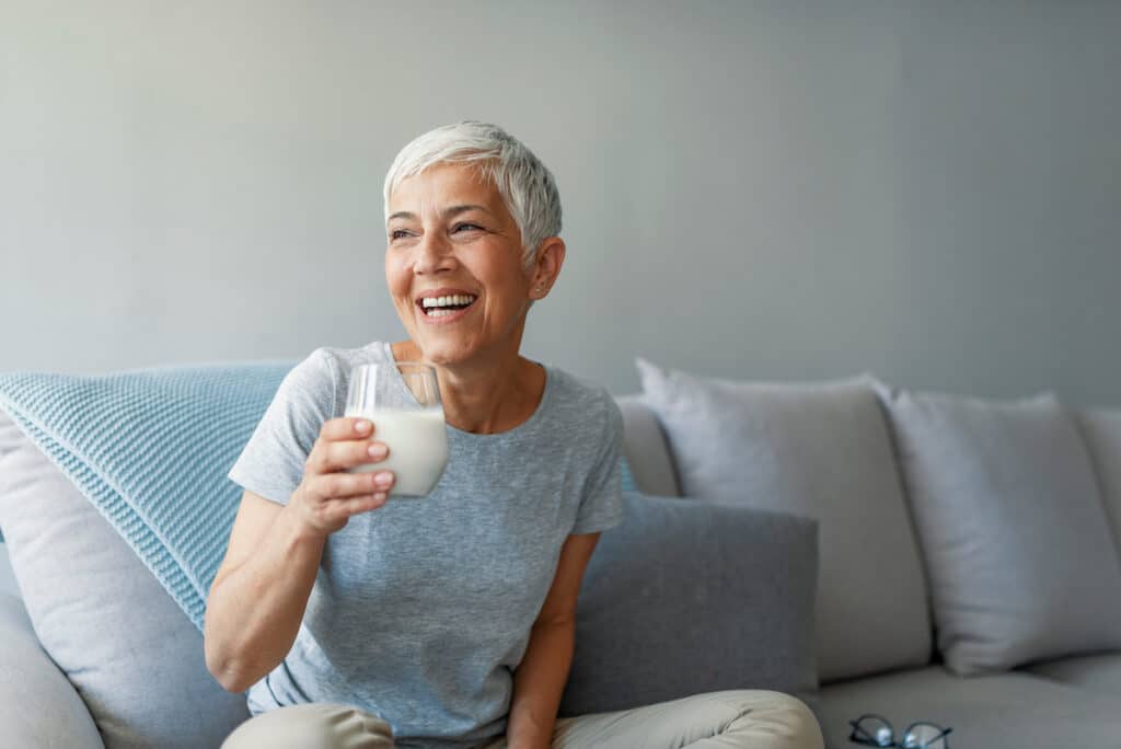 An older woman is sitting on her couch, holding a glass of milk and smiling.