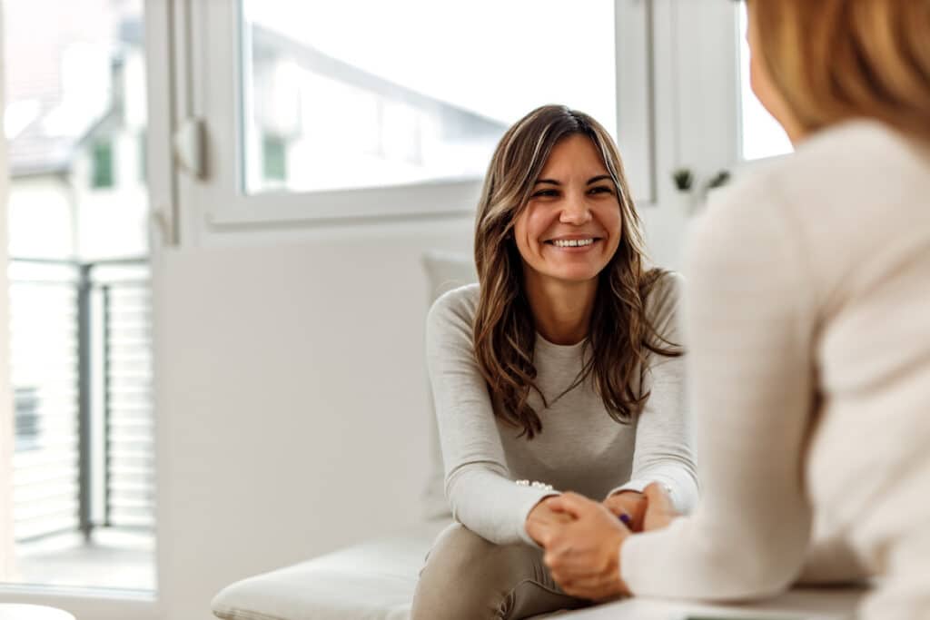 A woman is smiling at a behavioral health professional. They are sitting across from each other and the behavioral health professional is holding the patient's hands.