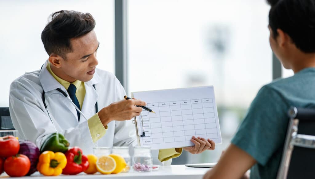 A positive doctor smiling and demonstrating a table to male patient while sitting at desk with healthy food. They are going over a meal plan.