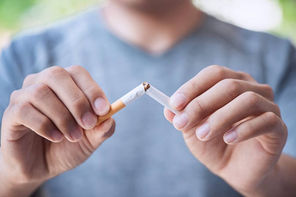 A close-up of a young adult male holding a cigarette in both hands. He's breaking the cigarette in half.