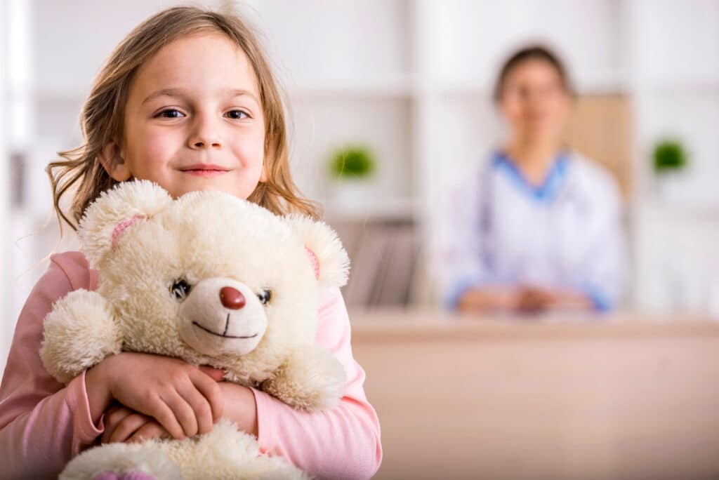 A little girl with a teddy bear is looking at the camera. Female doctor in the background.