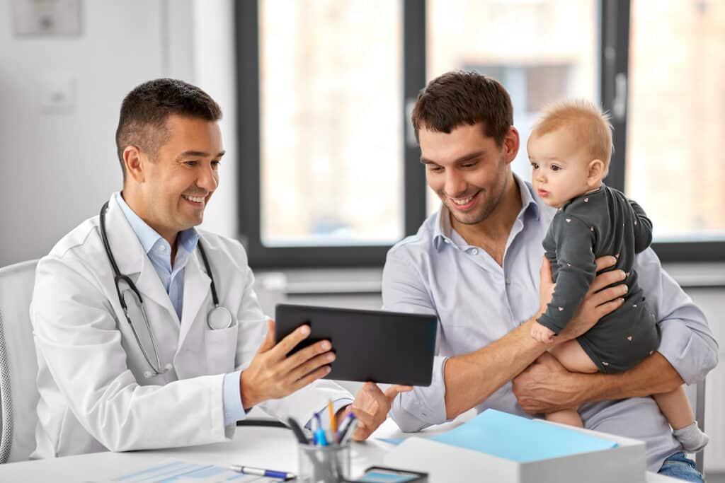 A man is holding his baby in the doctor's office. He's sitting at a table with the doctor, who is holding up a table to show the father something. They are both smiling.