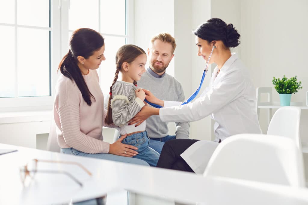 A mother and father are in an exam room with their young daughter. The female doctor is also there and has her stethoscope to the girl's chest, listening to her breathing.