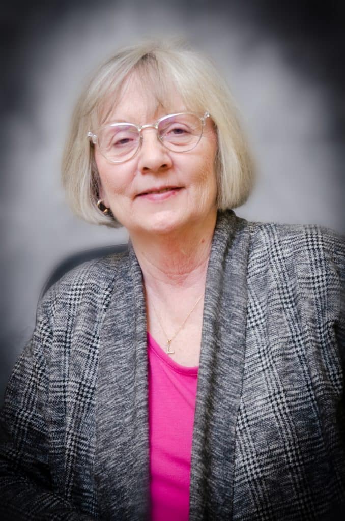 A headshot of Elizabeth Davenport. She is older, white, and has a short blonde bob haircut. She wears glasses and a pink shirt with a grey blazer over it.