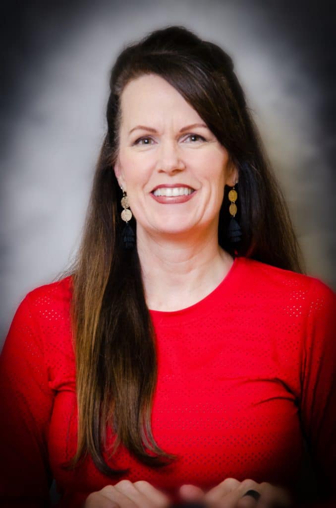 A headshot of Karen James. She's middle-aged, white, with long dark brown hair. She's wearing a red shirt.
