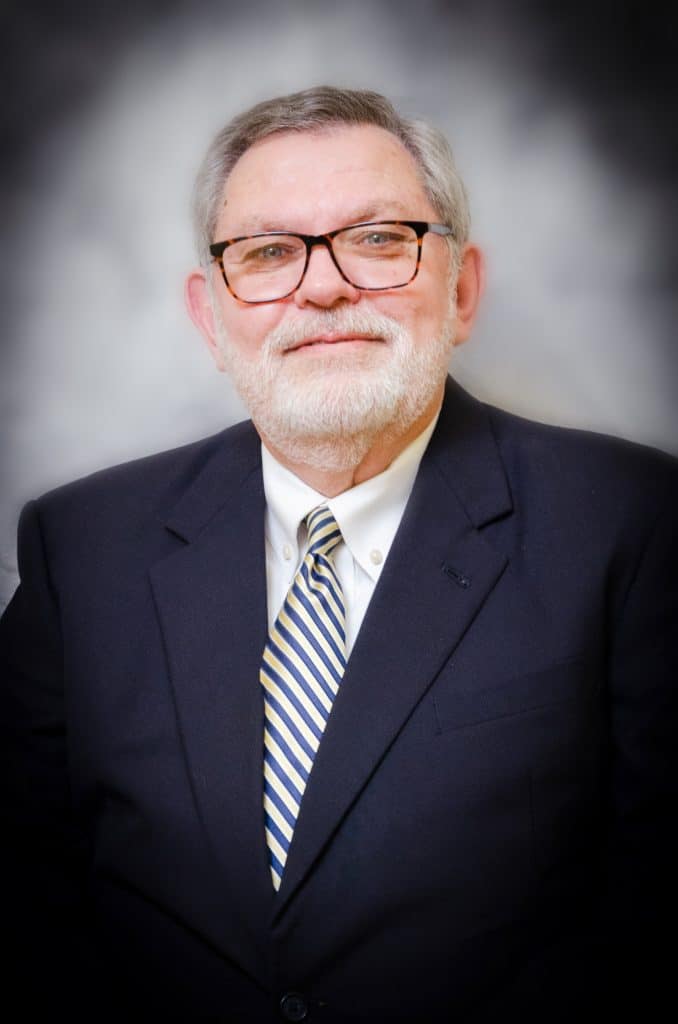 A headshot of Rick Glover. He's older, white, with short grey hair. He's wearing glasses, a white button up, black suit jacket, and a blue and yellow striped tie.